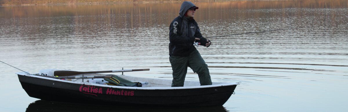Ein Angler ist auf einem Boot und angelt. Im Hintergrund ist der See & Bäume
