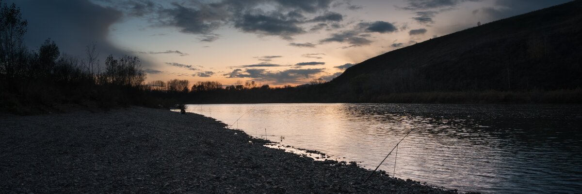 Landschaftsaufnahme eines Flusses. Am Ufer befinden sich Steine & Angelruten. Im Hintergrund geht die Sonne unter.