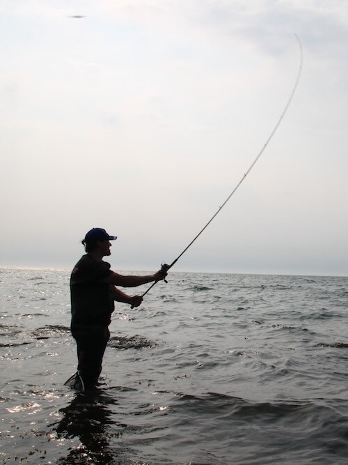 Ein Angler steht im Wasser und wirft den Köder mit seiner Angelrute aus. Ringsherum ist das Meer