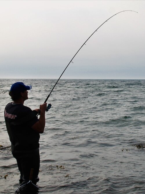 Ein Angler steht im Wasser mit seiner Angelrute und drillt einen Fisch. Ringsherum ist das Meer