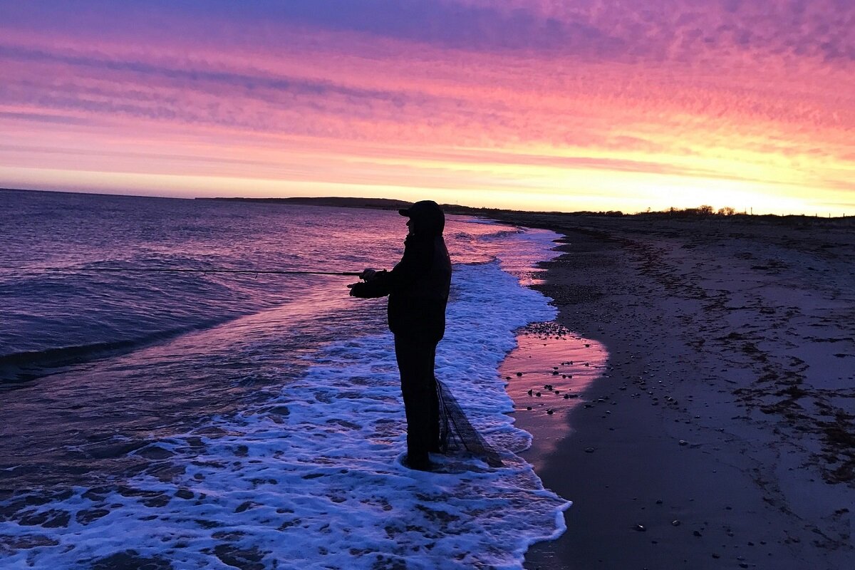 Angler steht am Strand zum Meer gerichtet & im Hintergrund ist der Sonnenaufgang.