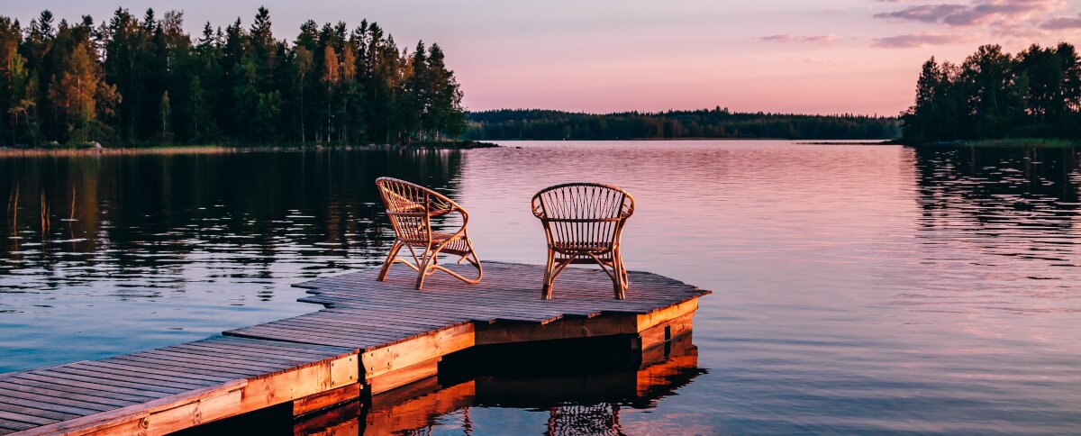 Aufnahme von zwei Stühlen, die auf einem Steg am See stehen. Abendröte im Hintergrund.