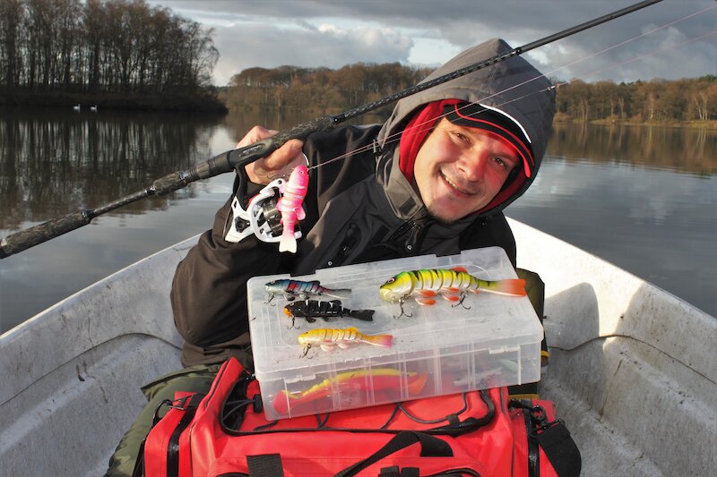 Im Vordergrund hält eine Angler auf einem Boot eine Angelrute, eine Angeltasche mit einer Box & Ködern. Im Hintergrund ist das Gewässer & Bäume.