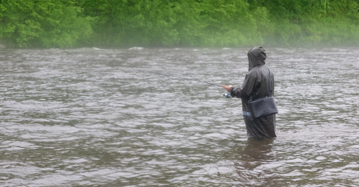 Aufnahme von einem Angler, der bei Regen mitten in einem Fluss steht und angelt.
