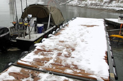 Im Vordergrund ein Steg mit Schnee. Daneben liegt ein Angelboot mit Angelruten & Zelt auf dem Wasser.