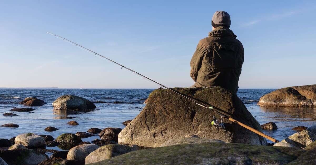 Im Vordergrund Angler, der ein einem Felsen lehnt. Im Hintergrund das Meer.
