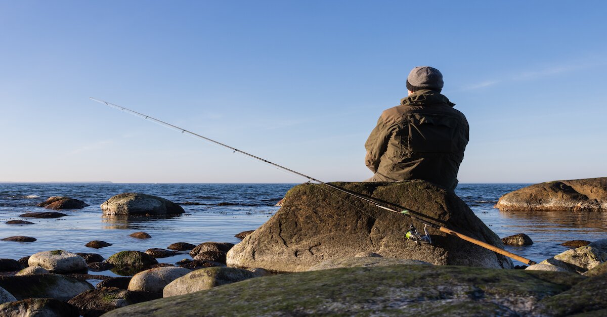 Aufnahme eines Anglers von hinten, sitzend auf einem Stein und aufs Meer blickend.