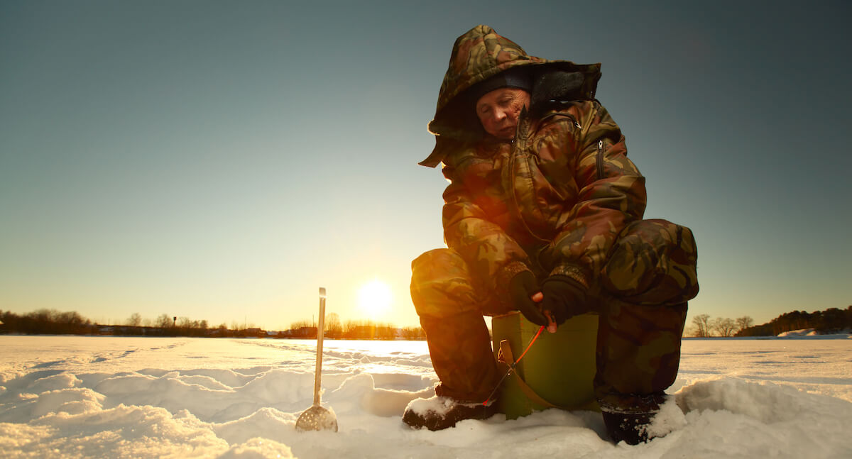 Angler in Thermoanzug am schneebedeckten See bei sonniger Winterzeit: 100%; height: 100%;